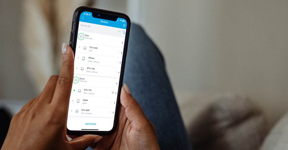 Photo of a person holding a phone showing the Orbi app's device management screen. It lists two people, each with three devices assigned to them.